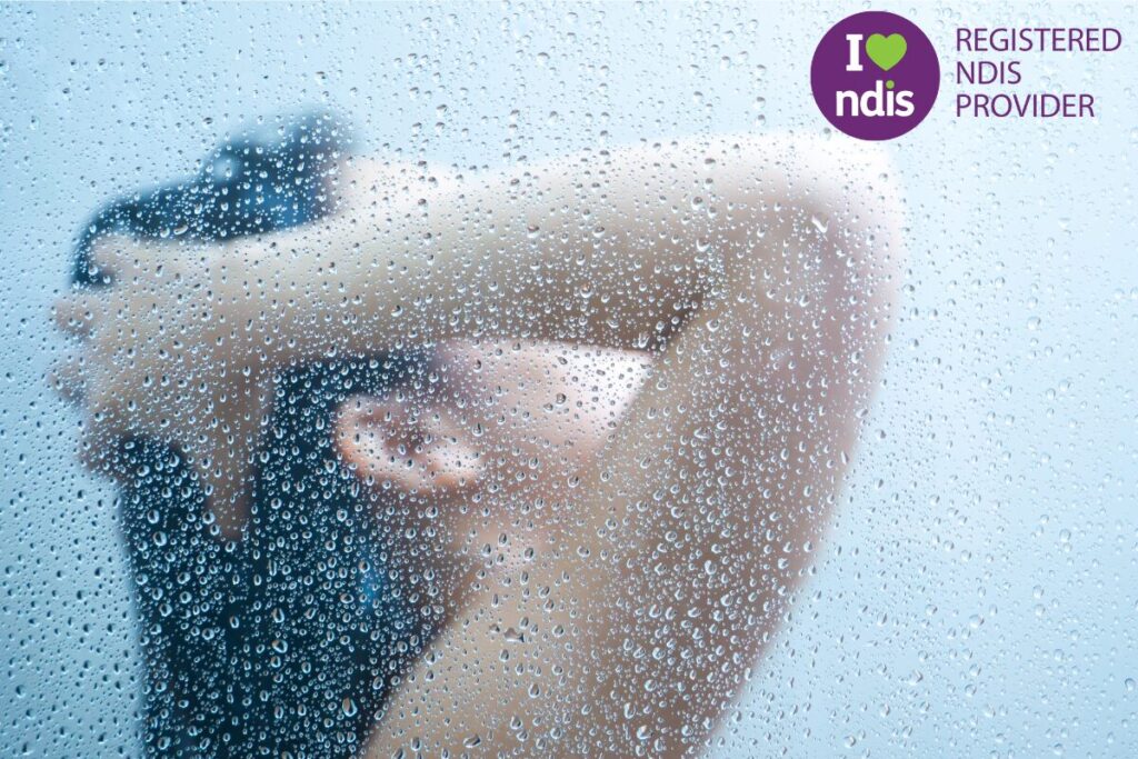 Close-up view of a person showering, with water droplets covering the glass screen and an NDIS Registered Provider logo in the corner.