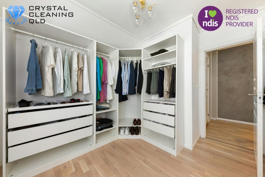 A clean and organised white walk-in wardrobe with neatly hung clothes and timber floors, featuring the Crystal Cleaning QLD logo and a Registered NDIS Provider badge.