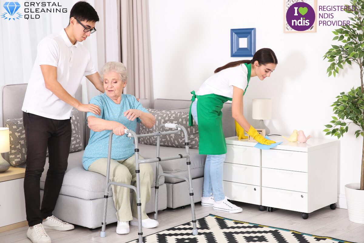 A young male support worker assists an elderly woman using a walker, while a female cleaner in a green apron dusts a cabinet nearby, as shown in image_11.png. The Crystal Cleaning QLD and Registered NDIS Provider logos are visible, illustrating how to perfect cleaning and support services easily.