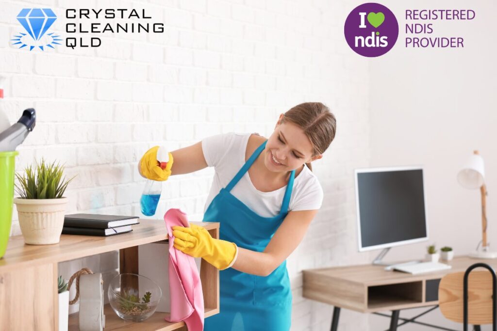 A smiling female cleaner wearing a blue apron and yellow gloves uses a spray bottle and cloth to wipe down wooden shelving in a bright office environment, as shown in 名称未設定 (1200 x 800 px) (12).jpg. The Crystal Cleaning QLD and Registered NDIS Provider logos are displayed in the upper corners to complement the guide on how you can perfect cleaning and support services easily.