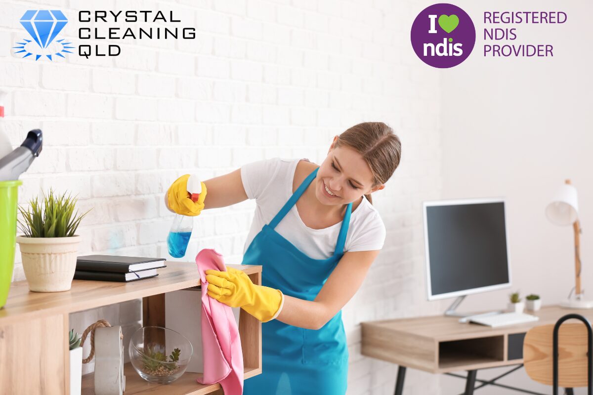 A smiling female cleaner wearing a blue apron and yellow gloves uses a spray bottle and cloth to wipe down wooden shelving in a bright office environment, as shown in 名称未設定 (1200 x 800 px) (12).jpg. The Crystal Cleaning QLD and Registered NDIS Provider logos are displayed in the upper corners to complement the guide on how you can perfect cleaning and support services easily.