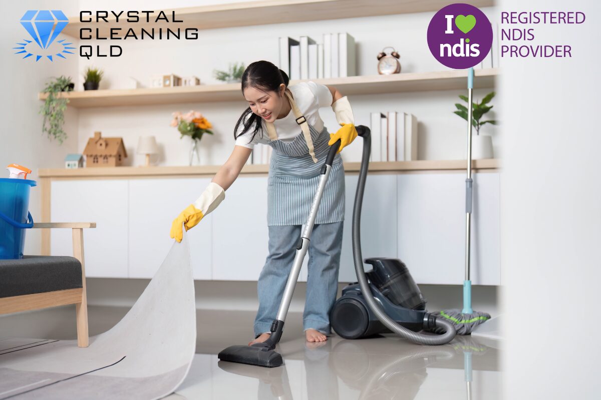 A smiling female cleaner wearing a striped apron and yellow gloves lifts a rug to vacuum underneath the tiled floor in a bright living area, as shown in 名称未設定 (1200 x 800 px) (13).jpg. The Crystal Cleaning QLD and Registered NDIS Provider logos are displayed in the upper corners to complement the guide on how to perfect cleaning and support services easily with staff training.