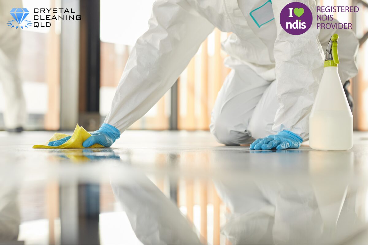 A professional cleaning technician in a white protective hazmat suit and blue gloves wiping a glossy floor with a yellow cloth next to a spray bottle, as shown in 名称未設定 (1200 x 800 px) (15).jpg. The Crystal Cleaning QLD and Registered NDIS Provider logos are displayed in the upper corners to complement the step-by-step infection control guide on how to perfect cleaning & support easily.