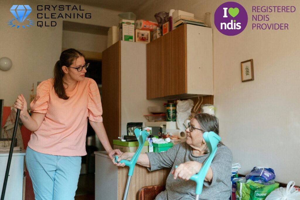 An support worker in a peach top interacting warmly with an elderly participant holding forearm crutches inside a home, as shown in 名称未設定 (1200 x 800 px) (17).jpg. The Crystal Cleaning QLD and Registered NDIS Provider logos are displayed in the upper corners to complement the article on how to perfect cleaning & support easily.