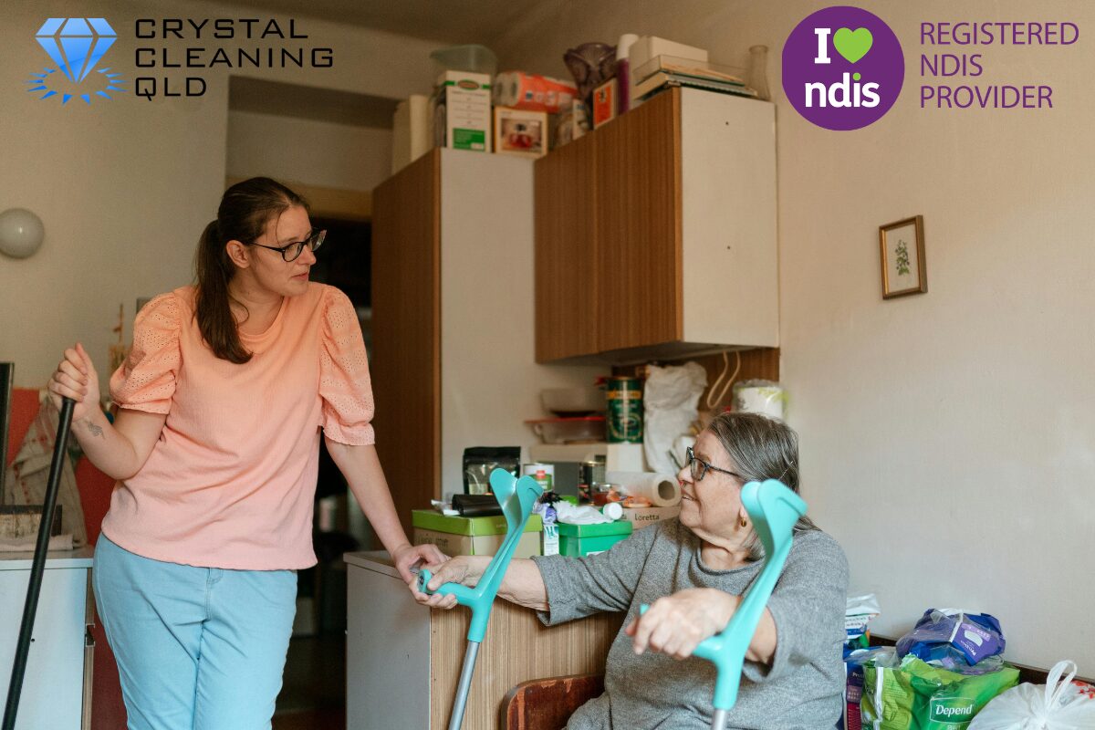 An support worker in a peach top interacting warmly with an elderly participant holding forearm crutches inside a home, as shown in 名称未設定 (1200 x 800 px) (17).jpg. The Crystal Cleaning QLD and Registered NDIS Provider logos are displayed in the upper corners to complement the article on how to perfect cleaning & support easily.
