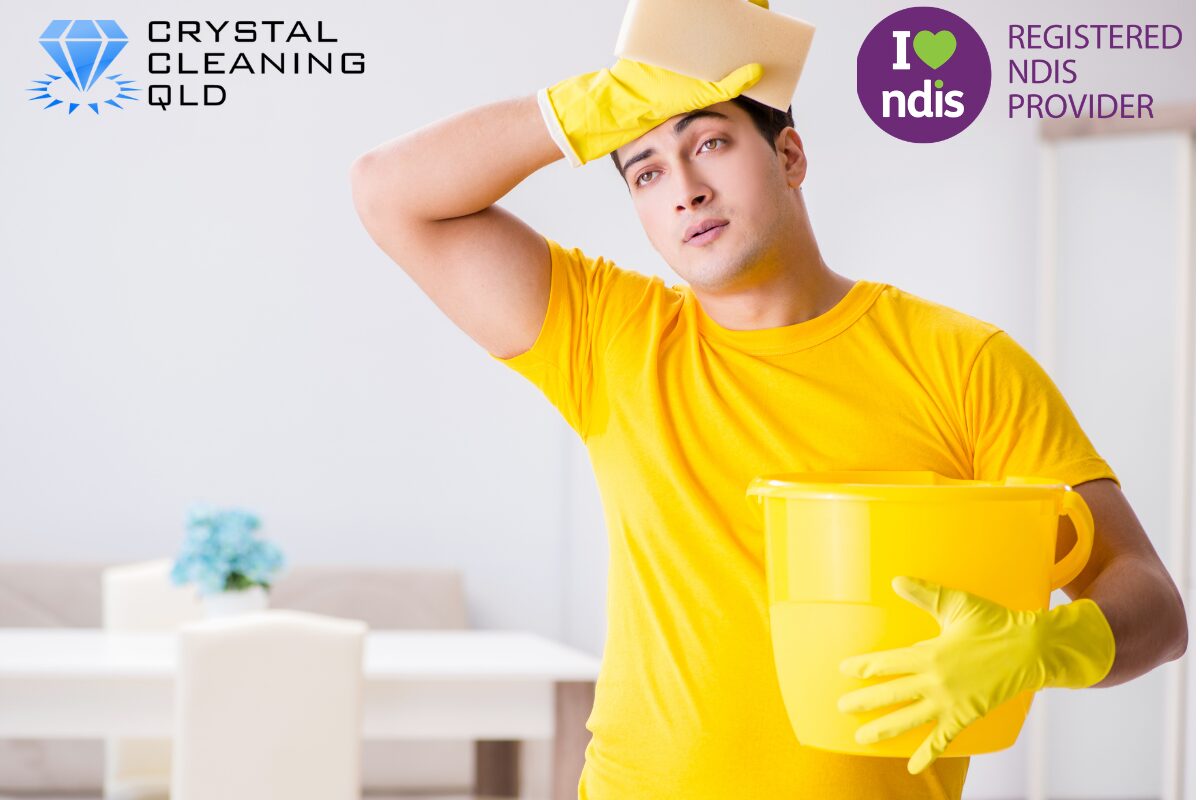 A tired cleaner in a yellow t-shirt and rubber gloves wiping sweat from his forehead with a sponge while carrying a yellow bucket in a bright home. The Crystal Cleaning QLD and Registered NDIS Provider logos are shown in the upper corners to highlight the challenges of house chores and how to perfect cleaning & support easily.