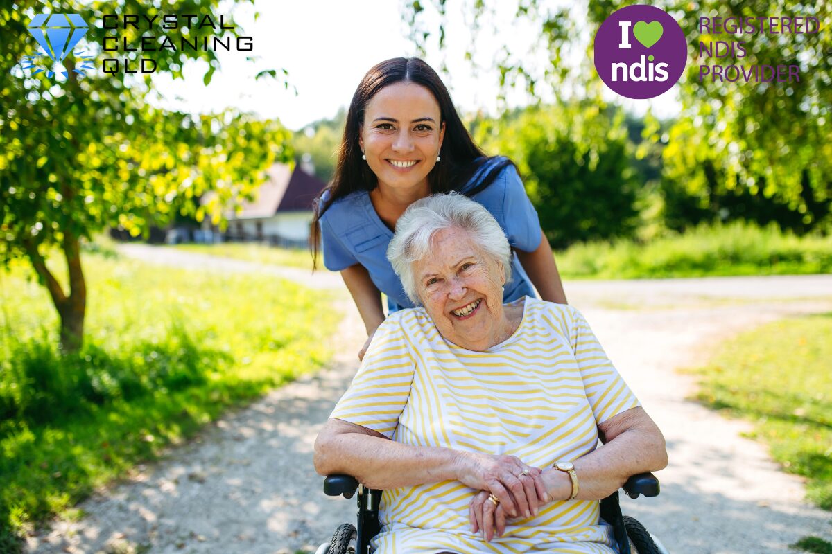 A smiling female support worker leaning over a senior woman in a wheelchair outdoors, featuring the Crystal Cleaning QLD and Registered NDIS Provider logos. This image illustrates how we understand resident needs for NDIS and aged care cleaning in Brisbane and the Gold Coast.