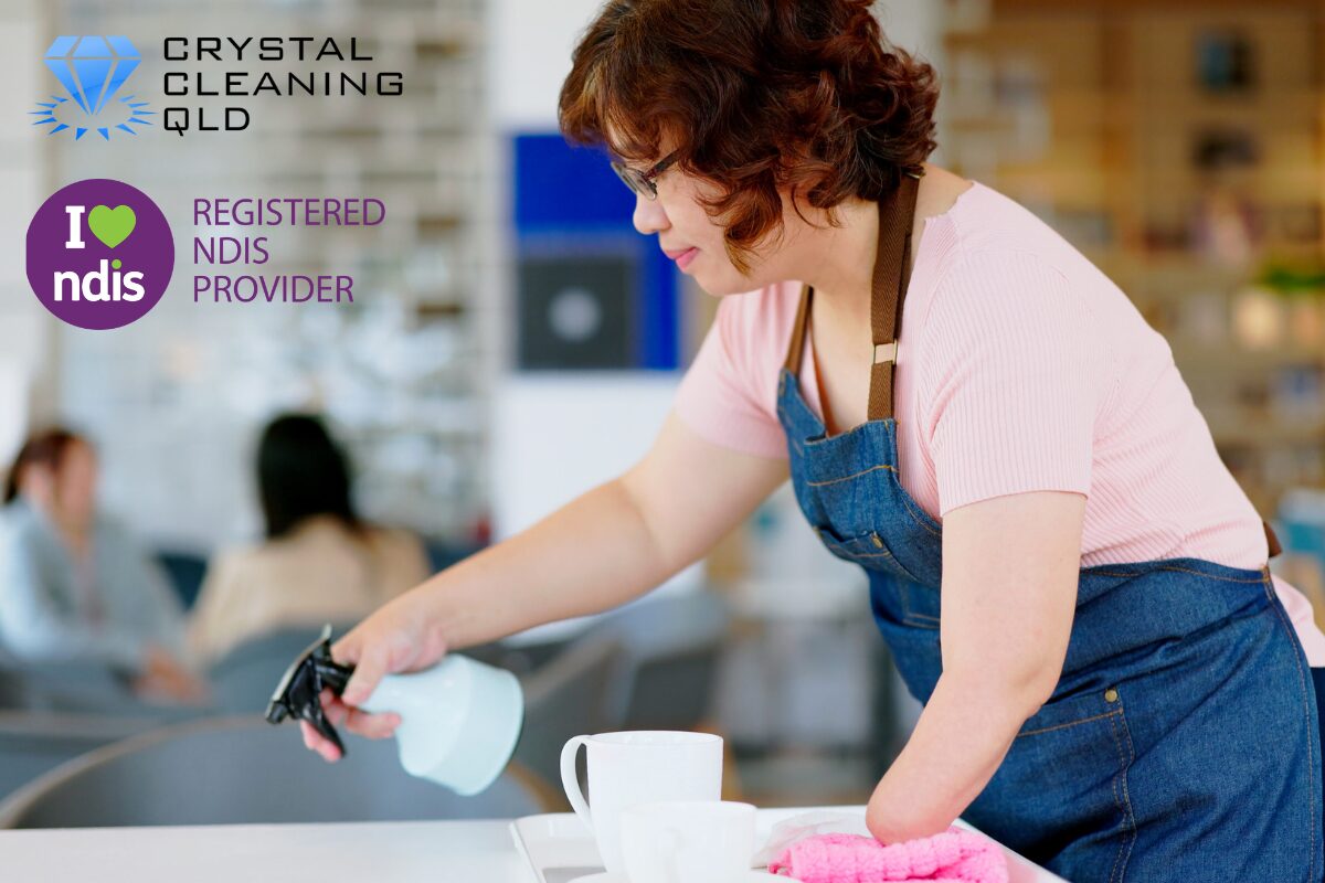 A professional support worker cleaning a tabletop surface with a spray bottle and cloth, featuring the Crystal Cleaning QLD and Registered NDIS Provider logos.