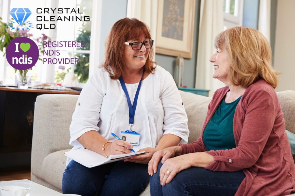 A support coordinator or support worker with a notepad consulting and smiling with an NDIS participant on a living room couch, featuring the Crystal Cleaning QLD and Registered NDIS Provider logos.