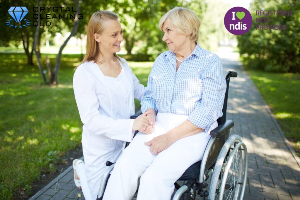 A friendly professional support worker holding hands and smiling with an elderly woman in a wheelchair outdoors, demonstrating compassionate NDIS funded support care.