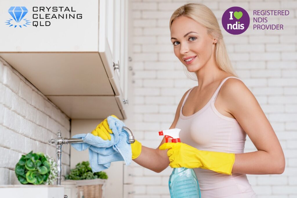 A professional female cleaner wearing yellow protective gloves, smiling while holding a spray bottle and wiping down a kitchen tap with a microfiber cloth, delivering compliant cleaning and support services easily.
