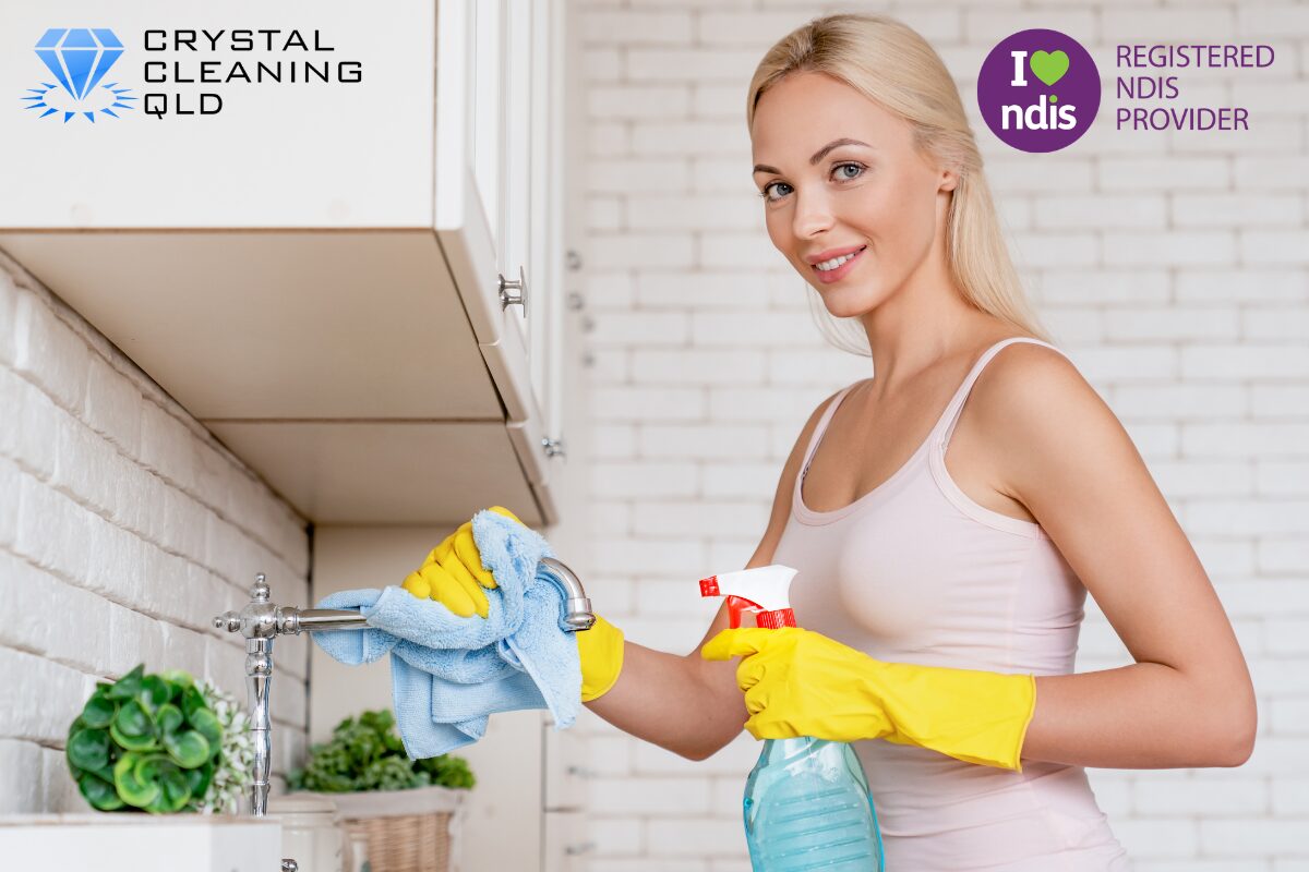 A professional female cleaner wearing yellow protective gloves, smiling while holding a spray bottle and wiping down a kitchen tap with a microfiber cloth, delivering compliant cleaning and support services easily.