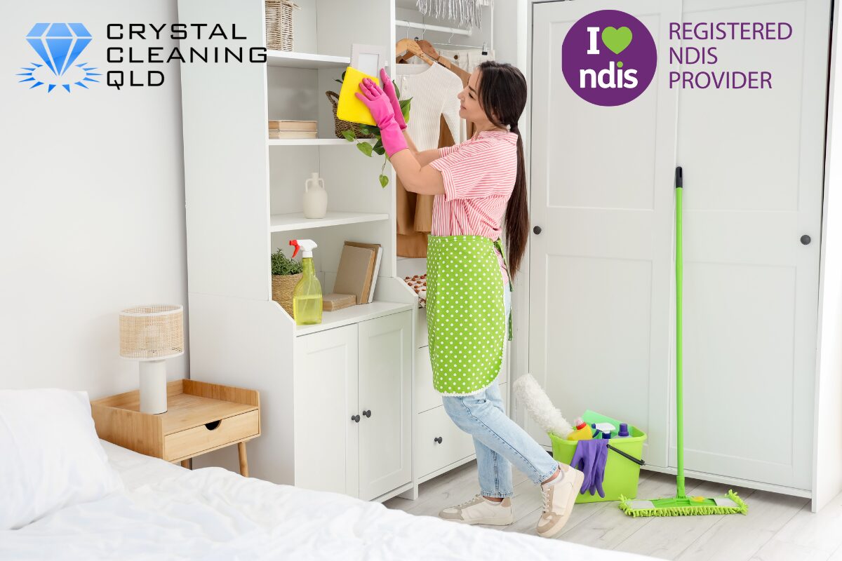 A professional woman from Crystal Cleaning QLD wearing pink gloves and a green apron while carefully dusting white bedroom shelves, demonstrating accessible home care and NDIS domestic assistance services in a tidy Brisbane property.