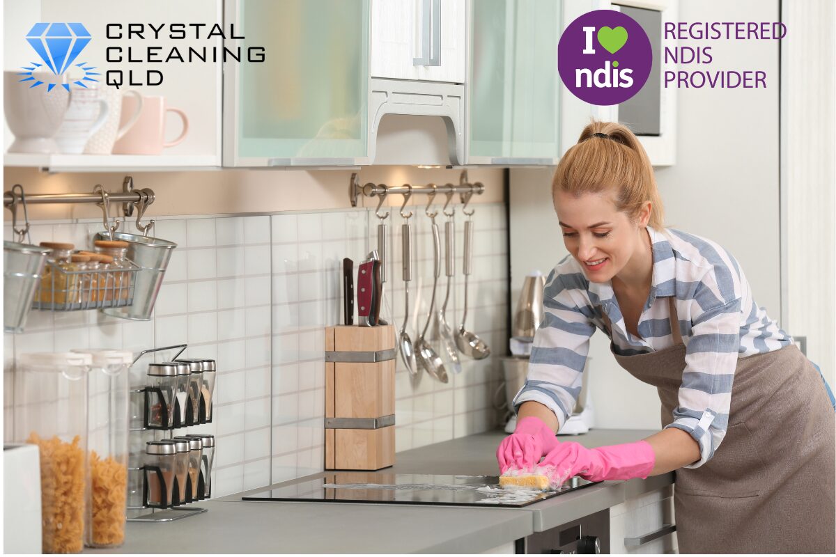 A professional woman from Crystal Cleaning QLD wearing pink gloves and an apron while smiling and carefully wiping down a glass cooktop in a bright, modern kitchen, highlighting safe domestic assistance services provided by an NDIS Registered Provider.